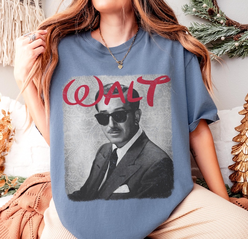 May include: A blue t-shirt featuring a black and white image of a man wearing sunglasses and a suit. The word "WALT" is written in red cursive above the image. The shirt has short sleeves.