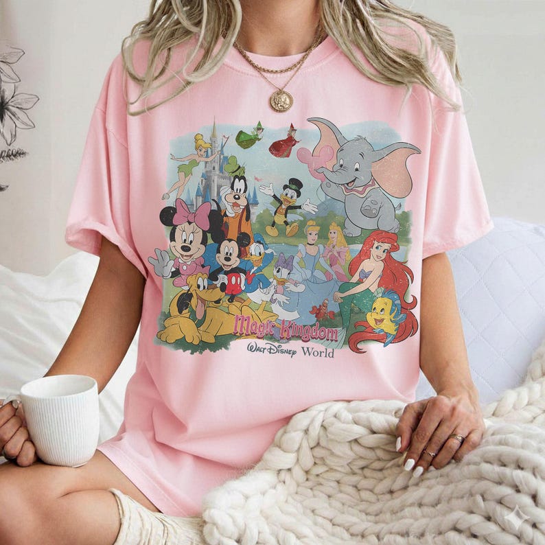 Puede incluir: Camiseta rosa claro con un gr&aacute;fico colorido de personajes de Disney, incluyendo Mickey y Minnie Mouse, Donald Duck y Dumbo. El texto "Magic Kingdom Walt Disney World" est&aacute; impreso debajo. La camiseta tiene mangas cortas.