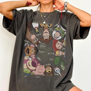 K&ouml;nnte beinhalten: Dunkelgraues T-Shirt mit einer farbenfrohen Grafik von Charakteren aus dem Animationsfilm "Toy Story". Das Design umfasst Woody, Buzz Lightyear, Jessie und andere Charaktere mit ihren Namen. Das T-Shirt hat einen Vintage-Look.
