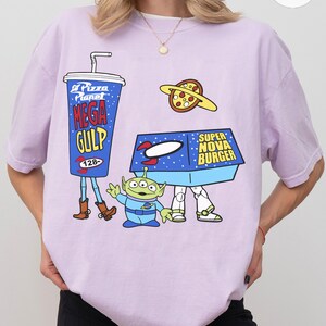 以下が含まれることがあります： トイ・ストーリーの漫画グラフィックが特徴の薄紫色のTシャツ。デザインには、メガガルフドリンク、ピザプラネットピザ、スーパーノヴァバーガーボックス、緑色のエイリアンキャラクターが含まれています。
