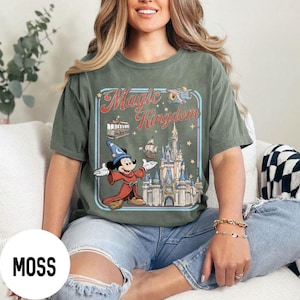 May include: Moss green t-shirt featuring a graphic of Mickey Mouse in a sorcerer's outfit, the Magic Kingdom logo, and a castle. The design is framed in a light blue rectangle. The shirt is a relaxed fit.