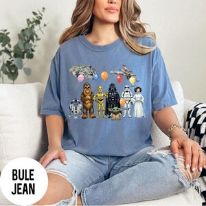 Peut inclure: Un t-shirt bleu avec des personnages et des &eacute;l&eacute;ments de Star Wars, dont le Faucon Millenium, un X-wing et divers personnages comme Dark Vador et Yoda. Le t-shirt porte le texte "BULE JEAN" en bas.