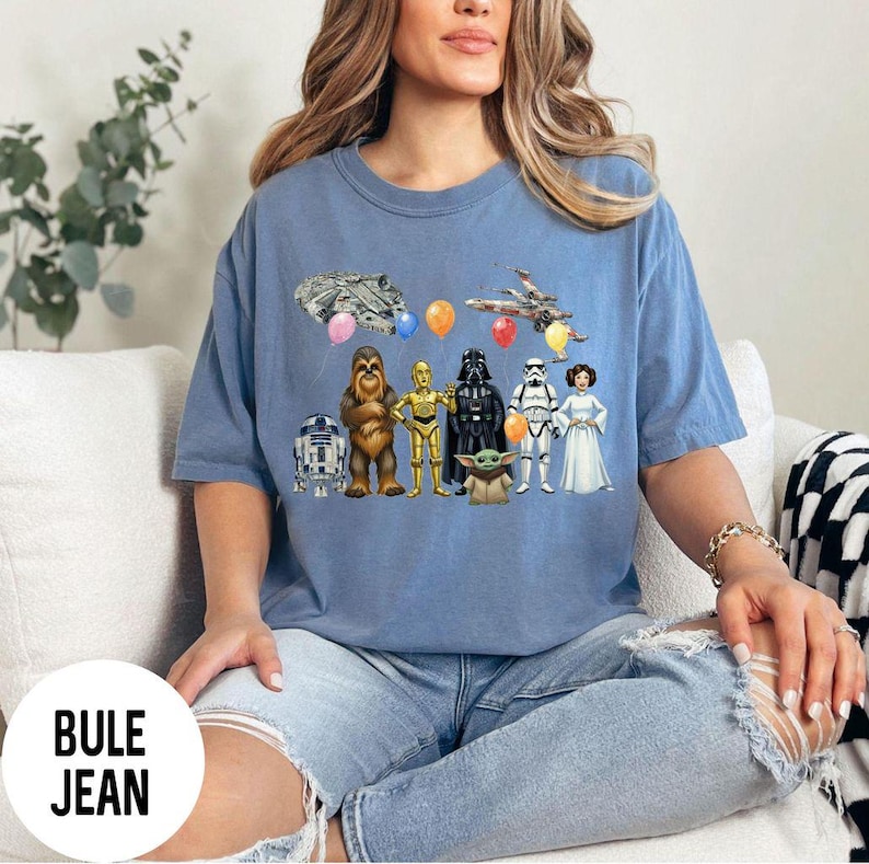 Peut inclure: Un t-shirt bleu avec des personnages et des &eacute;l&eacute;ments de Star Wars, dont le Faucon Millenium, un X-wing et divers personnages comme Dark Vador et Yoda. Le t-shirt porte le texte "BULE JEAN" en bas.
