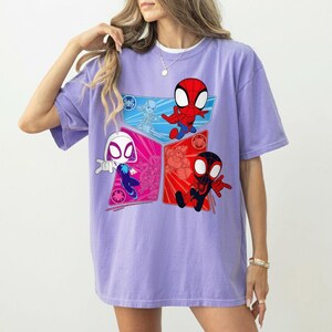 May include: A light purple t-shirt featuring a colorful graphic of Spider-Man characters. The design includes Spider-Man, Spider-Gwen, and Miles Morales in a cartoon style. The shirt has short sleeves and a relaxed fit.