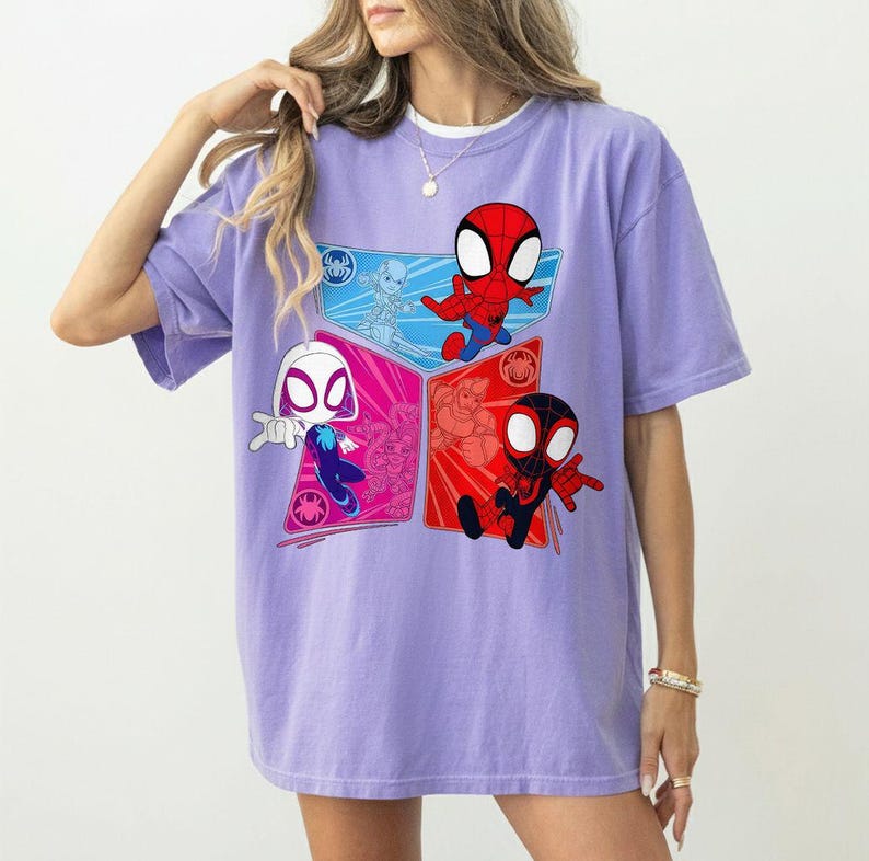 May include: A light purple t-shirt featuring a colorful graphic of Spider-Man characters. The design includes Spider-Man, Spider-Gwen, and Miles Morales in a cartoon style. The shirt has short sleeves and a relaxed fit.