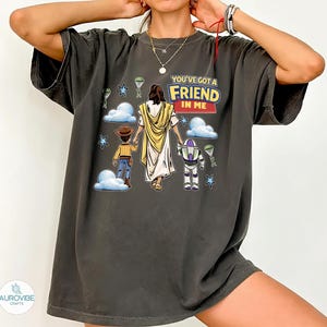 May include: Dark gray t-shirt featuring a graphic of Jesus, Woody, and Buzz Lightyear with the text "YOU'VE GOT A FRIEND IN ME." The design includes clouds, stars, and parachutes. The shirt has short sleeves and a relaxed fit.