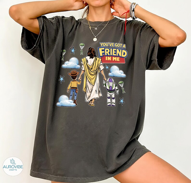 May include: Dark gray t-shirt featuring a graphic of Jesus, Woody, and Buzz Lightyear with the text "YOU'VE GOT A FRIEND IN ME." The design includes clouds, stars, and parachutes. The shirt has short sleeves and a relaxed fit.