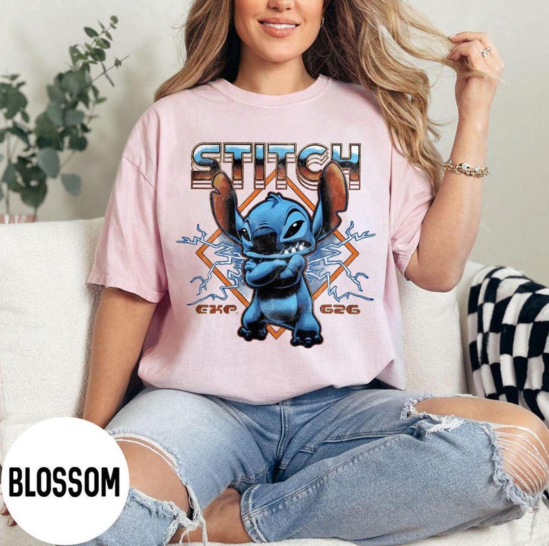 Puede incluir: Camiseta rosa claro con un personaje de dibujos animados azul, orejas grandes y la palabra "STITCH" en una fuente retro. El personaje est&aacute; sobre un dise&ntilde;o en forma de diamante con rayos y el texto "G26" y "GKP".