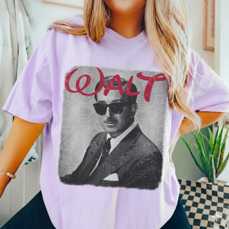 May include: A light purple t-shirt featuring a black and white image of a man wearing sunglasses and a suit. The word "WALT" is written in red cursive above the image. The shirt is a casual style.