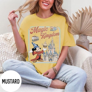 May include: Mustard-colored t-shirt featuring a graphic of Mickey Mouse in a sorcerer's outfit, the Magic Kingdom logo, and a castle. The design includes a trolley, Dumbo, and a ship. The shirt is a relaxed fit.