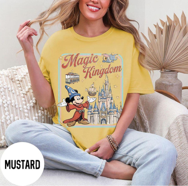 May include: Mustard-colored t-shirt featuring a graphic of Mickey Mouse in a sorcerer's outfit, the Magic Kingdom logo, and a castle. The design includes a trolley, Dumbo, and a ship. The shirt is a relaxed fit.