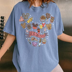 May include: A blue t-shirt featuring a graphic print of various snacks and treats arranged in a large Mickey Mouse head shape. The center of the design includes a pink banner with the text "BEST DAY EVER!" surrounded by small white stars.