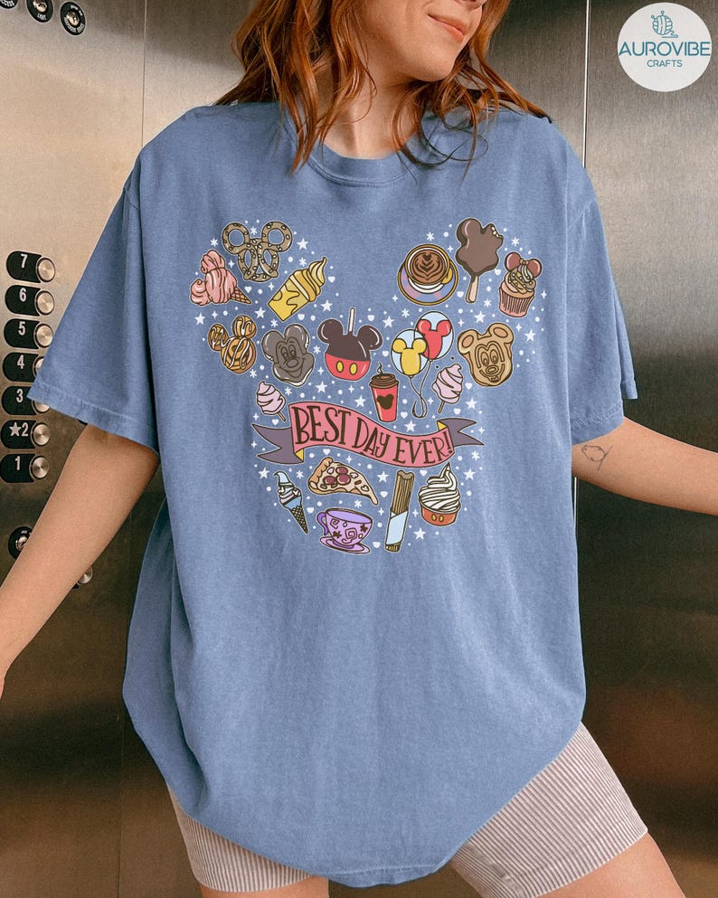 May include: A blue t-shirt featuring a graphic print of various snacks and treats arranged in a large Mickey Mouse head shape. The center of the design includes a pink banner with the text "BEST DAY EVER!" surrounded by small white stars.