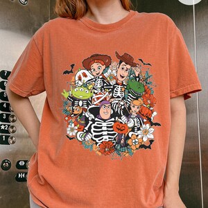 May include: A rust-colored short-sleeve t-shirt featuring a graphic print of various cartoon characters wearing skeleton costumes. The design includes spiderwebs, pumpkins, and flowers, with characters like a space ranger and a cowboy.