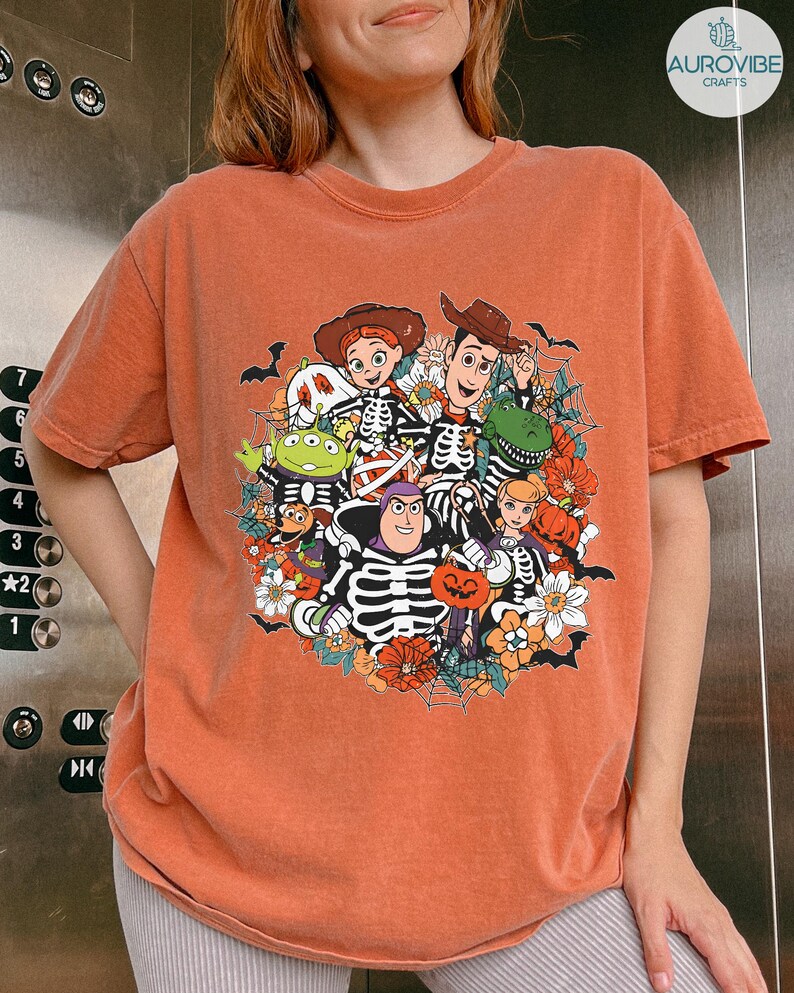 May include: A rust-colored short-sleeve t-shirt featuring a graphic print of various cartoon characters wearing skeleton costumes. The design includes spiderwebs, pumpkins, and flowers, with characters like a space ranger and a cowboy.