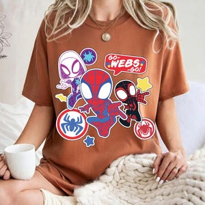 Puede incluir: Una camiseta marr&oacute;n con un gr&aacute;fico colorido de personajes de Spider-Man. El dise&ntilde;o incluye a Spider-Man, Spider-Gwen y Miles Morales, con bocadillos que dicen "Go Webs Go!" y emblemas de ara&ntilde;a.