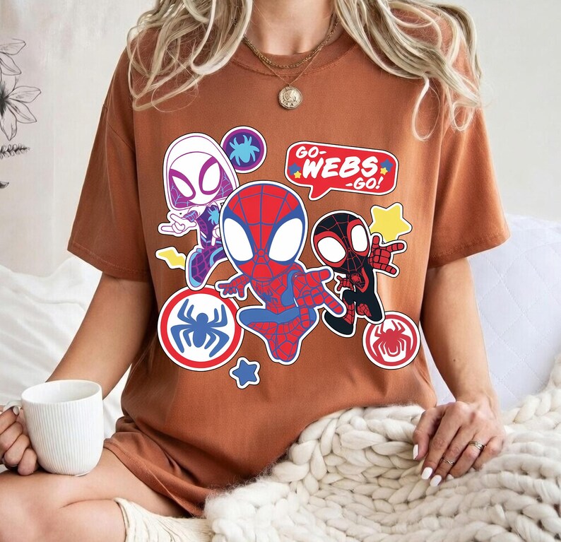 Puede incluir: Una camiseta marr&oacute;n con un gr&aacute;fico colorido de personajes de Spider-Man. El dise&ntilde;o incluye a Spider-Man, Spider-Gwen y Miles Morales, con bocadillos que dicen "Go Webs Go!" y emblemas de ara&ntilde;a.
