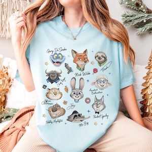 Op de afbeelding: Lichtblauw T-shirt met cartoonkarakterportretten en namen. Personages zijn onder meer Nick Wilde, Judy Hopps en Flash. Het shirt heeft een ronde hals en korte mouwen.