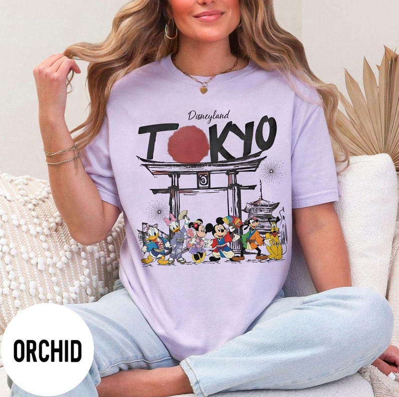 Puede incluir: Camiseta color orqu&iacute;dea con un gr&aacute;fico de Disneyland Tokyo. El dise&ntilde;o presenta la palabra "Tokyo" en letras negras grandes, un sol rojo y personajes de dibujos animados. La camiseta es de color morado claro.