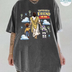 May include: Dark gray t-shirt featuring a graphic of Jesus holding hands with two cartoon characters, a cowboy and a space ranger. The text "YOU'VE GOT A FRIEND IN ME" is above the figures. Parachutes and stars are also in the design.