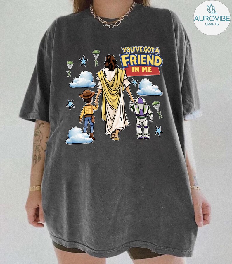 May include: Dark gray t-shirt featuring a graphic of Jesus holding hands with two cartoon characters, a cowboy and a space ranger. The text "YOU'VE GOT A FRIEND IN ME" is above the figures. Parachutes and stars are also in the design.