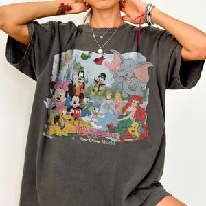 Puede incluir: Camiseta gris oscuro con un gr&aacute;fico colorido de personajes de Disney, incluyendo Mickey Mouse, Minnie Mouse, Donald Duck y Dumbo. El gr&aacute;fico incluye el texto "Magic Kingdom Walt Disney World".