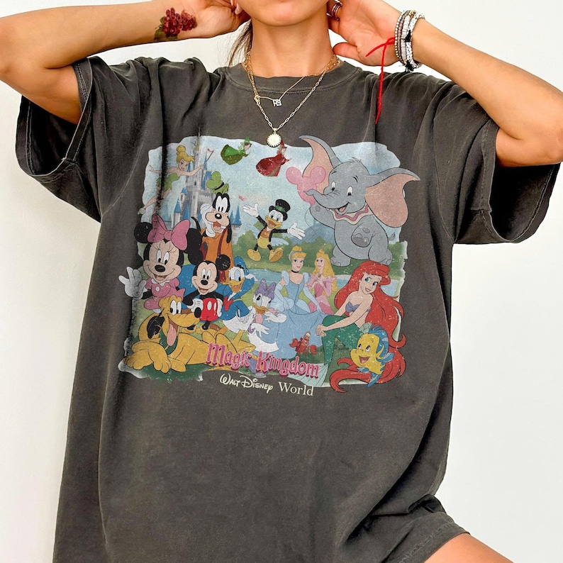 Puede incluir: Camiseta gris oscuro con un gr&aacute;fico colorido de personajes de Disney, incluyendo Mickey Mouse, Minnie Mouse, Donald Duck y Dumbo. El gr&aacute;fico incluye el texto "Magic Kingdom Walt Disney World".