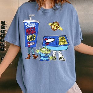 以下が含まれることがあります： 映画トイ・ストーリーの漫画グラフィックが特徴の青いTシャツ。デザインには、メガガルのカップ、ピザプラネット、スーパーノヴァバーガーボックス、緑色のエイリアンキャラクターが含まれています。シャツは半袖です。