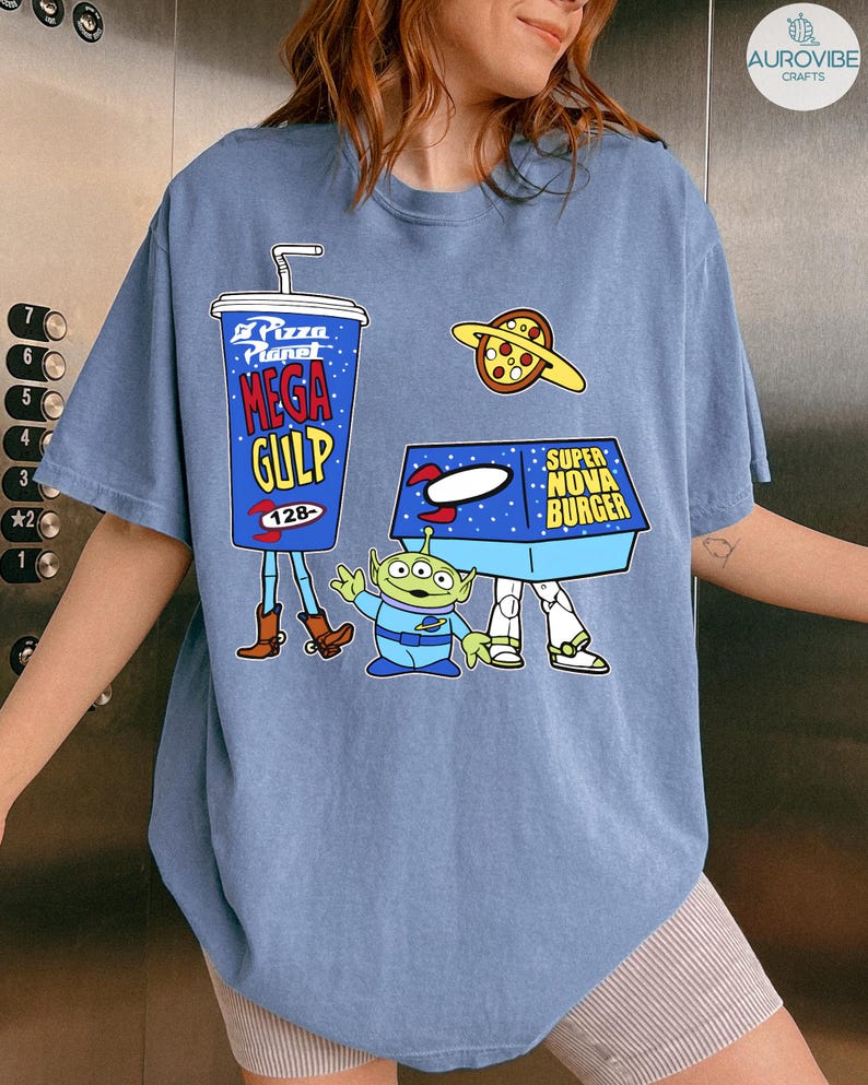 以下が含まれることがあります： 映画トイ・ストーリーの漫画グラフィックが特徴の青いTシャツ。デザインには、メガガルのカップ、ピザプラネット、スーパーノヴァバーガーボックス、緑色のエイリアンキャラクターが含まれています。シャツは半袖です。