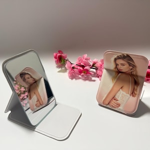 May include: A white and silver folding mirror and a matching phone stand, both featuring a woman in a white lace top. Pink flowers are in the background. The mirror is open and the phone stand is upright.