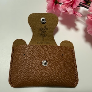 Könnte beinhalten: Ein brauner Kartenhalter aus strukturiertem Leder mit Druckknopfverschluss. Die Innenklappe zeigt eine gravierte Strichzeichnung einer Rose und den Text your text. Der Kartenhalter hat zwei kleine Löcher für Ohrringe und eine abgerundete Klappe.