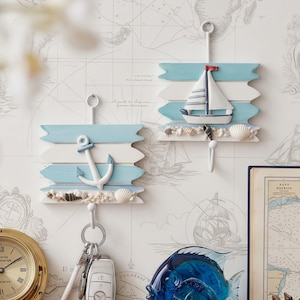 May include: Two nautical-themed wall hooks. One features a white anchor on blue and white wooden planks, the other a sailboat. Both have seashell accents and a metal hook for hanging keys or other items.