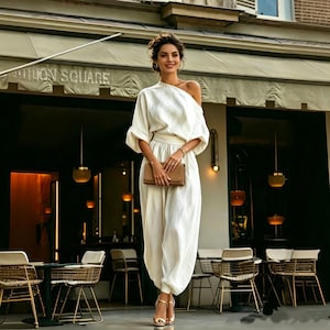May include: A woman in a white one-shoulder jumpsuit, matching belt, and gold heels, holding a brown clutch. The background features a restaurant with outdoor seating.