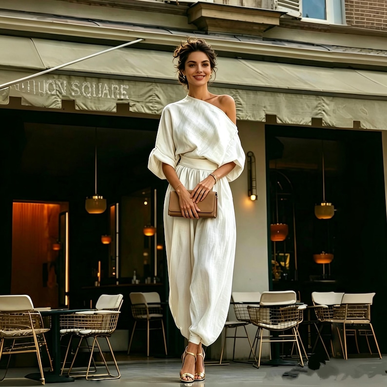 May include: A woman in a white one-shoulder jumpsuit, matching belt, and gold heels, holding a brown clutch. The background features a restaurant with outdoor seating.
