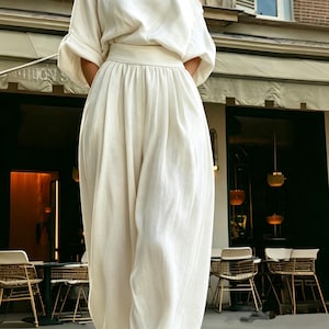 May include: A cream linen outfit, comprising a loose top with elbow-length sleeves and high-waisted, wide-leg trousers. The outfit has a relaxed, flowing silhouette. A restaurant with outdoor seating is visible in the background.