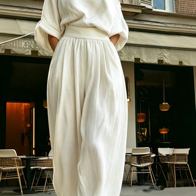 May include: A cream linen outfit, comprising a loose top with elbow-length sleeves and high-waisted, wide-leg trousers. The outfit has a relaxed, flowing silhouette. A restaurant with outdoor seating is visible in the background.