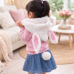 以下が含まれることがあります： 白いとピンクのウサギの耳のフードとデニムスカートを着た子供。フードは、ピンクの内側の耳が付いたふわふわの白い素材です。白いポンポンの尾がスカートに付いています。子供はピンクのスウェットシャツを着ています。