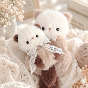 May include: Two plush stuffed animals, a larger one holding a smaller one, both featuring cream-coloured fur on their heads and dark brown fur on their bodies and ears. Each plush animal has black bead eyes and a white lace ribbon tied around its neck.