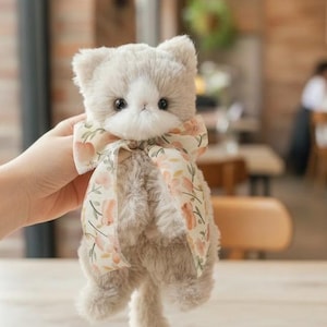 Puede incluir: Un peluche gris con forma de gato, con cara blanca y ojos negros grandes. Tiene un lazo con estampado floral. El gato está sostenido en una mano.