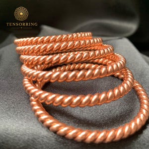 May include: A stack of five circular copper-colored rings with a twisted rope-like texture. Each ring features a continuous spiral pattern. The logo TENSORRING is visible in the upper left corner with a circular geometric icon above the text.