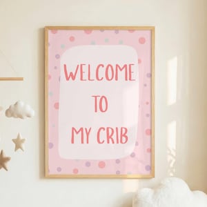 May include: A framed wall art print featuring a pink background with multicolored polka dots and the text WELCOME TO MY CRIB in coral pink letters. The print is displayed above a white crib with a hanging mobile of stars and clouds.