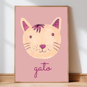 May include: A framed watercolor print featuring a cat's face in shades of beige and purple on a pink background. The word "gato" is written in cursive below the cat illustration. The artwork is displayed on a wooden floor.