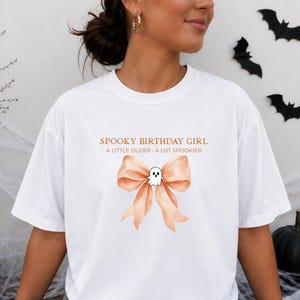 Peut inclure: Un t-shirt blanc avec un motif repr&eacute;sentant un n&oelig;ud en ruban couleur p&ecirc;che et un petit fant&ocirc;me blanc au centre. Le texte au-dessus du n&oelig;ud indique SPOOKY BIRTHDAY GIRL et A LITTLE OLDER - A LOT SPOOKIER dans une police orange simple.