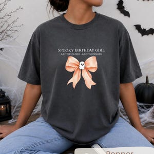 Peut inclure: Un t-shirt &agrave; col rond gris anthracite avec le texte SPOOKY BIRTHDAY GIRL A LITTLE OLDER A LOT SPOOKIER au-dessus d'un grand n&oelig;ud p&ecirc;che orn&eacute; d'une petite illustration de fant&ocirc;me blanc centr&eacute;e sur le n&oelig;ud du ruban.