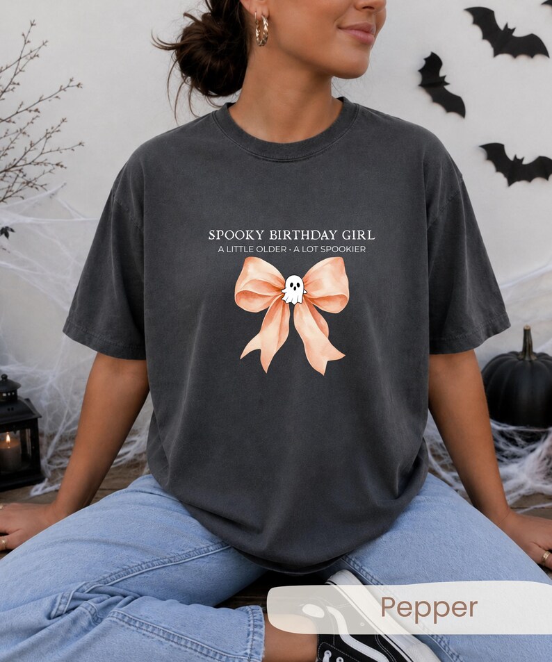 Peut inclure: Un t-shirt &agrave; col rond gris anthracite avec le texte SPOOKY BIRTHDAY GIRL A LITTLE OLDER A LOT SPOOKIER au-dessus d'un grand n&oelig;ud p&ecirc;che orn&eacute; d'une petite illustration de fant&ocirc;me blanc centr&eacute;e sur le n&oelig;ud du ruban.