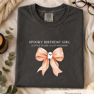 Peut inclure: Un t-shirt gris anthracite avec le texte blanc SPOOKY BIRTHDAY GIRL A LITTLE OLDER A LOT SPOOKIER au-dessus d'un grand n&oelig;ud p&ecirc;che clair orn&eacute; d'une petite illustration de fant&ocirc;me blanc plac&eacute;e au centre du n&oelig;ud.