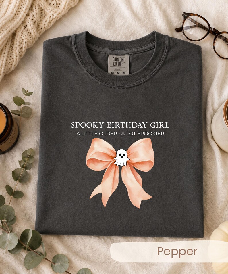 Peut inclure: Un t-shirt gris anthracite avec le texte blanc SPOOKY BIRTHDAY GIRL A LITTLE OLDER A LOT SPOOKIER au-dessus d'un grand n&oelig;ud p&ecirc;che clair orn&eacute; d'une petite illustration de fant&ocirc;me blanc plac&eacute;e au centre du n&oelig;ud.