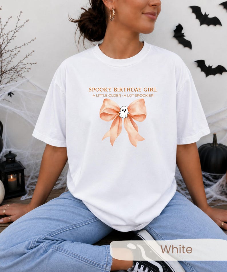 Peut inclure: Un t-shirt blanc avec un motif repr&eacute;sentant un n&oelig;ud en ruban couleur p&ecirc;che et un petit fant&ocirc;me blanc au centre. Le texte au-dessus du n&oelig;ud indique SPOOKY BIRTHDAY GIRL et A LITTLE OLDER - A LOT SPOOKIER dans une police orange simple.