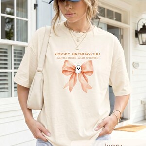 Peut inclure: Un t-shirt ivoire avec un motif repr&eacute;sentant un grand n&oelig;ud couleur p&ecirc;che et un petit fant&ocirc;me blanc au centre. Le texte au-dessus du motif indique SPOOKY BIRTHDAY GIRL et A LITTLE OLDER A LOT SPOOKIER. Le t-shirt a des manches courtes et un col rond.