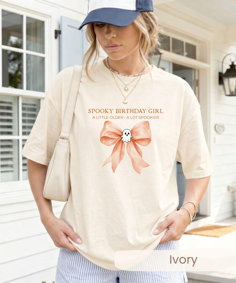 Peut inclure: Un t-shirt ivoire avec un motif repr&eacute;sentant un grand n&oelig;ud couleur p&ecirc;che et un petit fant&ocirc;me blanc au centre. Le texte au-dessus du motif indique SPOOKY BIRTHDAY GIRL et A LITTLE OLDER A LOT SPOOKIER. Le t-shirt a des manches courtes et un col rond.